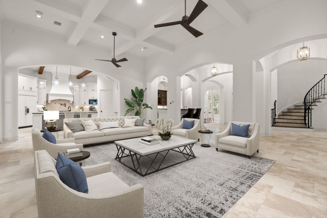 | the mckellar group Interior View of a Bright White Neutral Toned Living Room in a Luxurious High End Home Contains High Ceiling with Fan and a Couch and Chairs Around a Coffee Table on a Large Rug