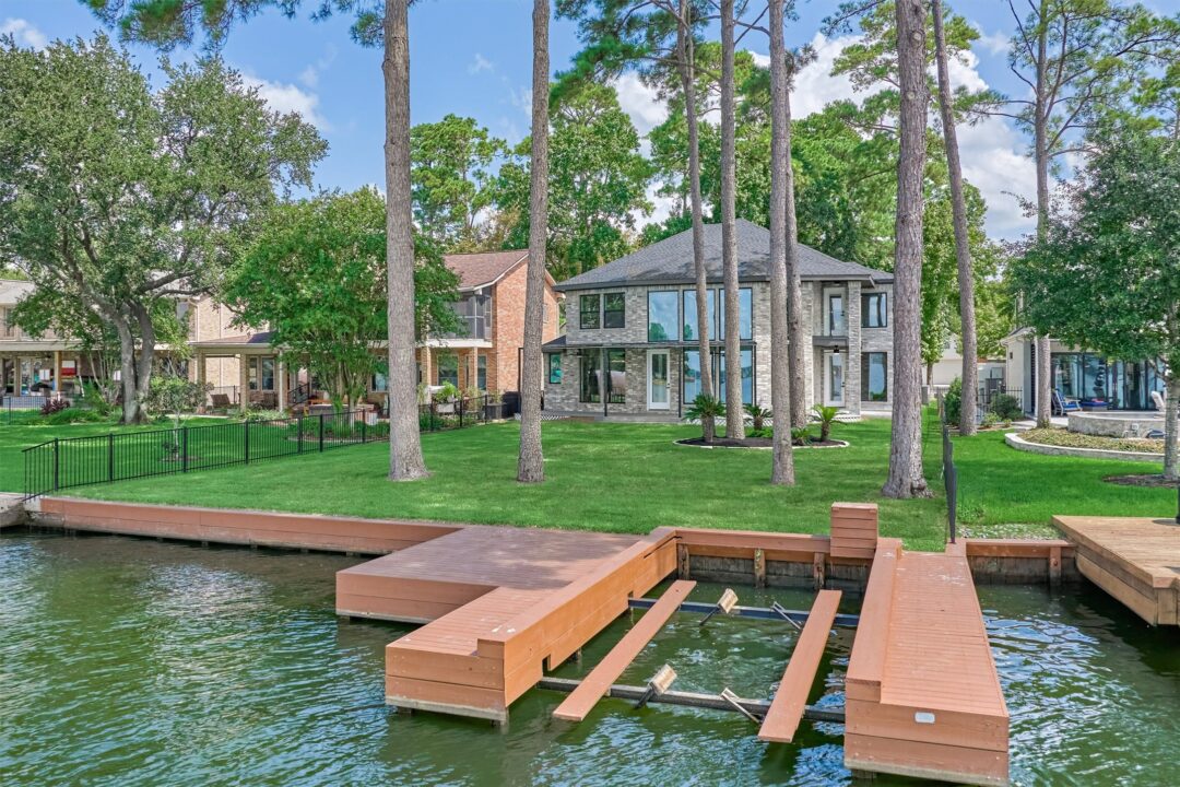 Modern Two story Gray brick House on a Green Lawn by a Canal with Tall Pine Trees and a Wooden Dock Extending into the Water