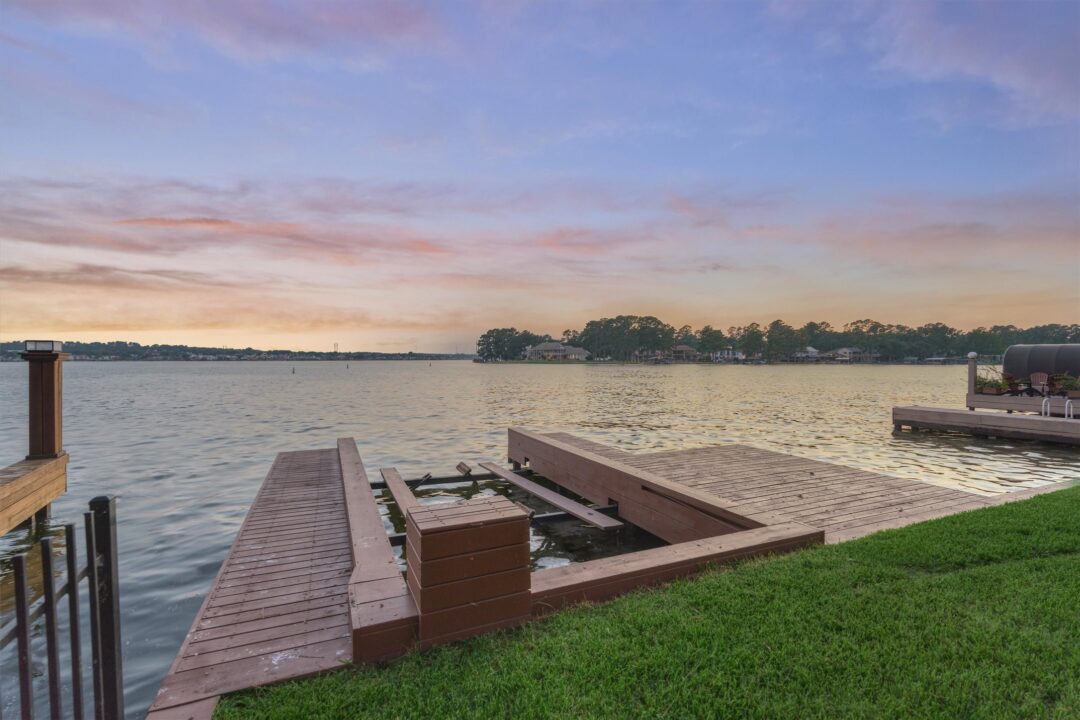 Waterfront buyer guide overview lake conroe home buying | the mckellar group Sunset over a Calm Lake with a Wooden Dock Extending from the Grassy Shore a Damaged Central Section with Broken Planks Sits Near the Water