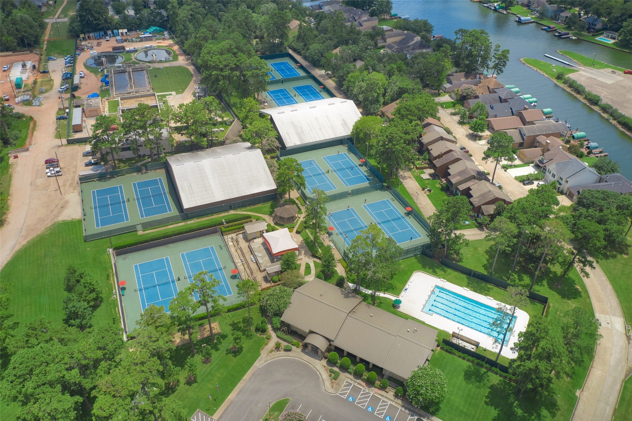 | the mckellar group Aerial View of a Sports Complex with Several Blue Tennis Courts Large White roofed Courts and Surrounding Green Fields Adjacent to Houses and a Canal