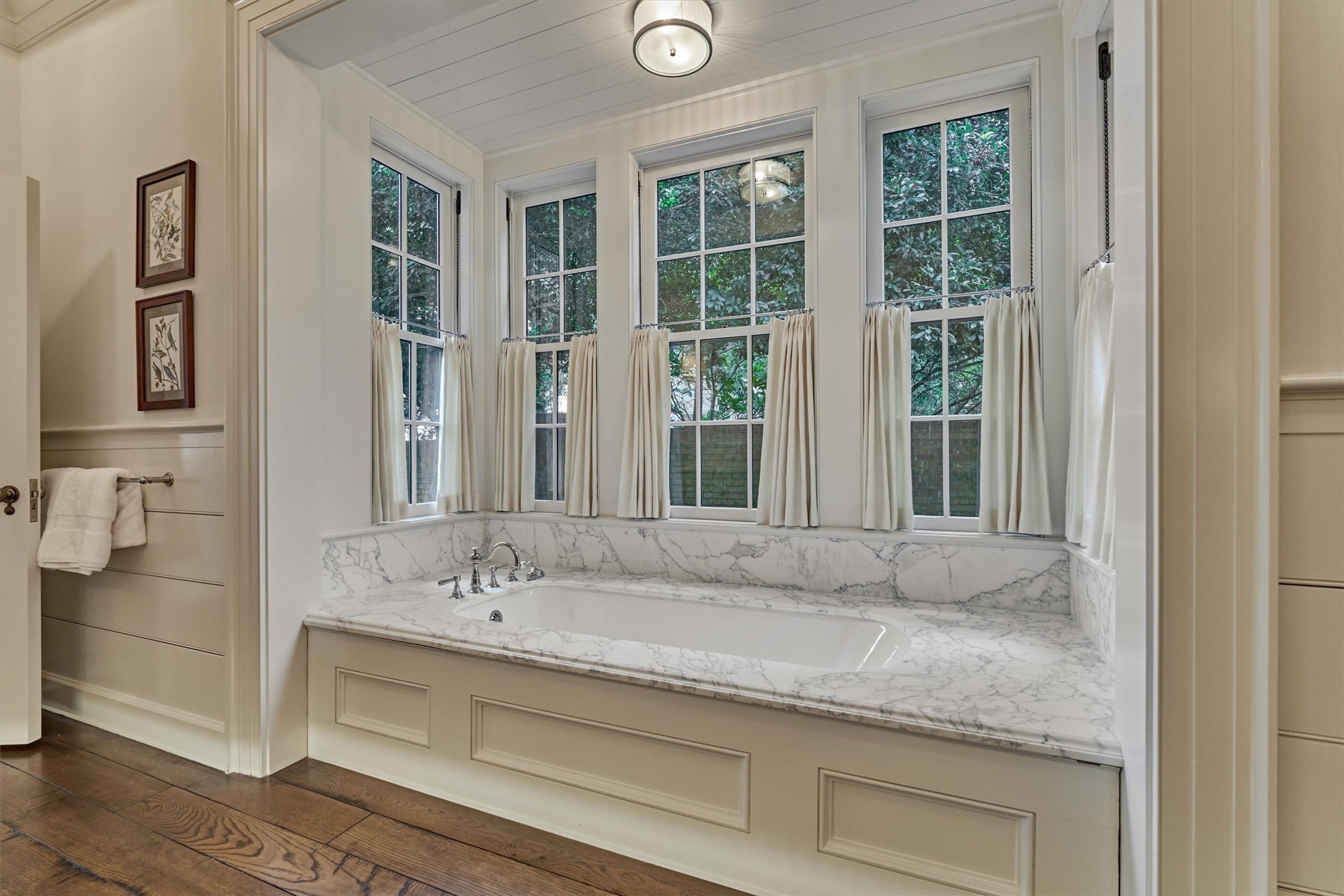 | the mckellar group Bathtub with a Marble Surround Beneath Tall Multi pane Windows Dressed with Cream Curtains in a Bright Traditional Bathroom
