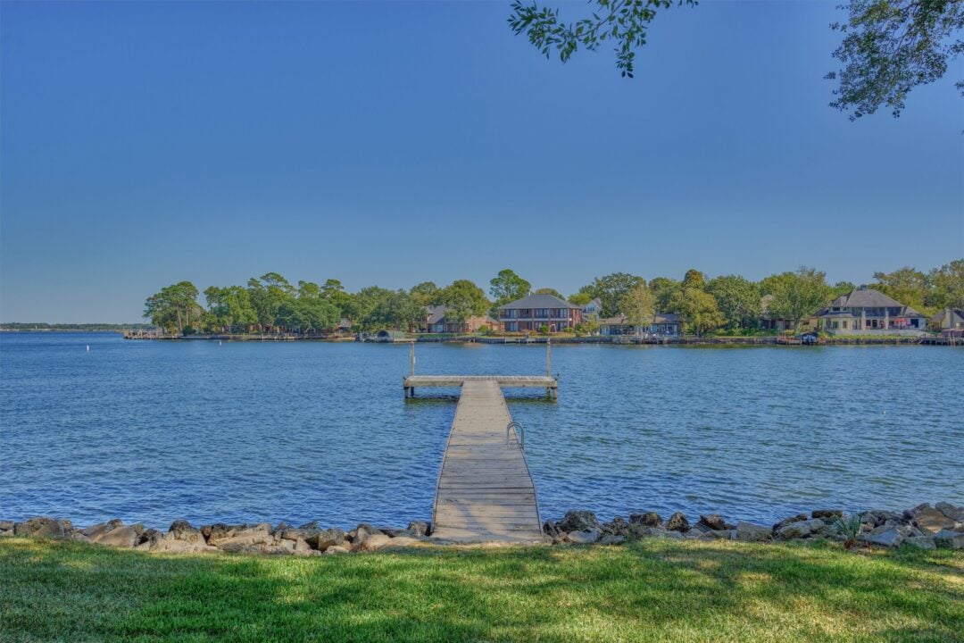 Long Wooden Pier Extends from Rocky Shore Toward Calm Lake with Houses and Trees Along the Far Shore Under a Clear Blue Sky