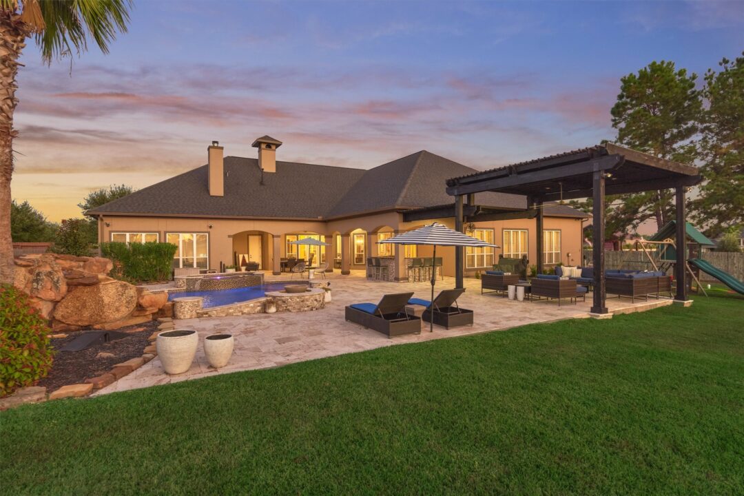 Backyard at Sunset with a Beige House Stone Patio and a Rectangular Pool Lounge Chairs Umbrellas and a Shaded Seating Area Under a Dark Pergola