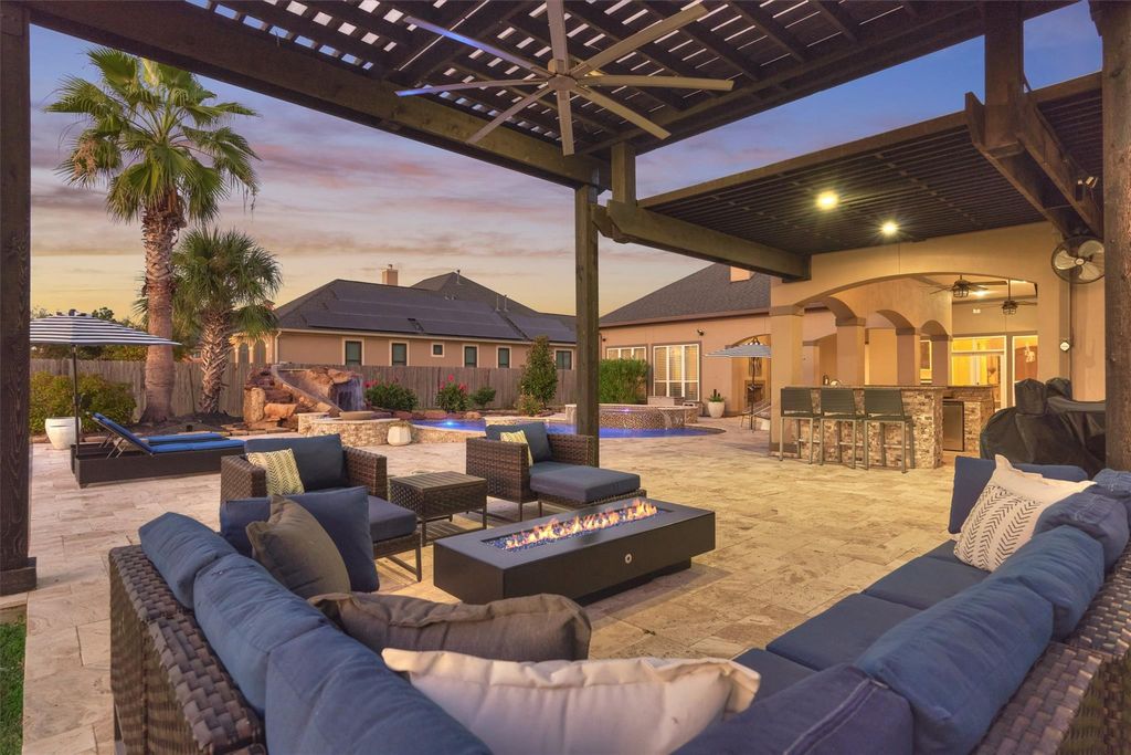 755979936 | the mckellar group Luxurious Backyard Lounge Under a Wooden Pergola with a Rectangular Gas Fire Pit Blue Cushioned Sofas Palm Trees and a Pool at Dusk