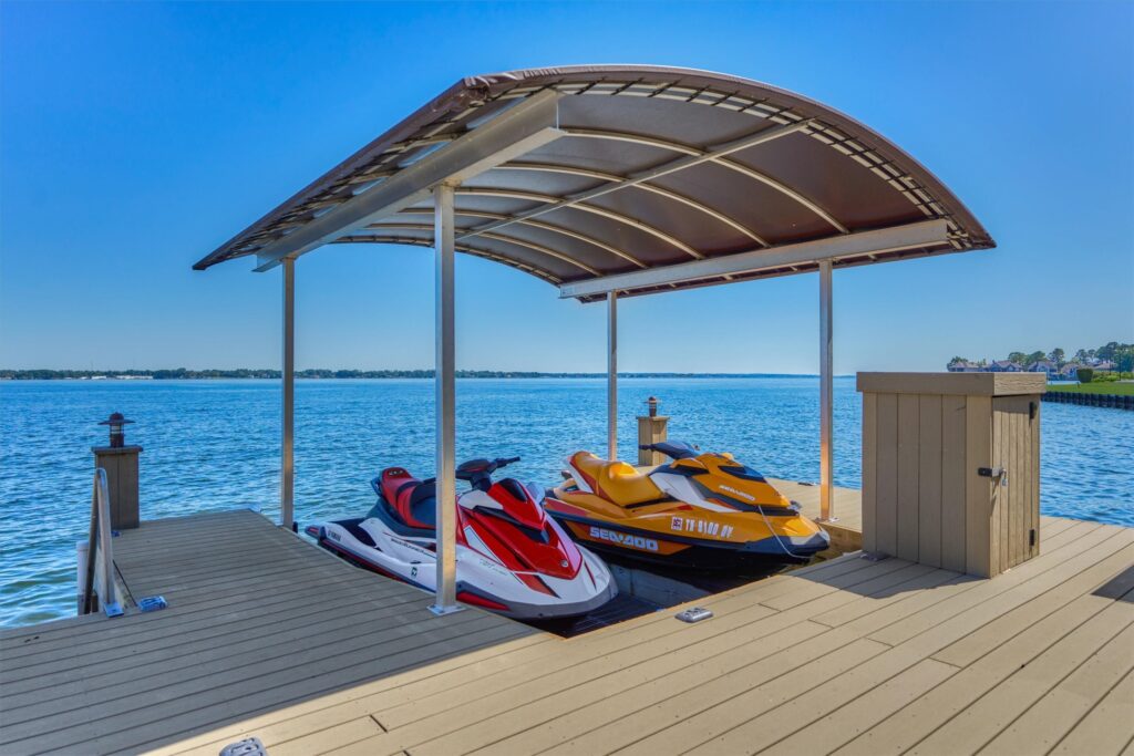 W 13 048 048 100 bentwater bay dr | the mckellar group Two Jet Skis Docked Under a Curved Canopy on a Wooden Pier Beside a Calm Blue Lake with a Distant Shore Visible