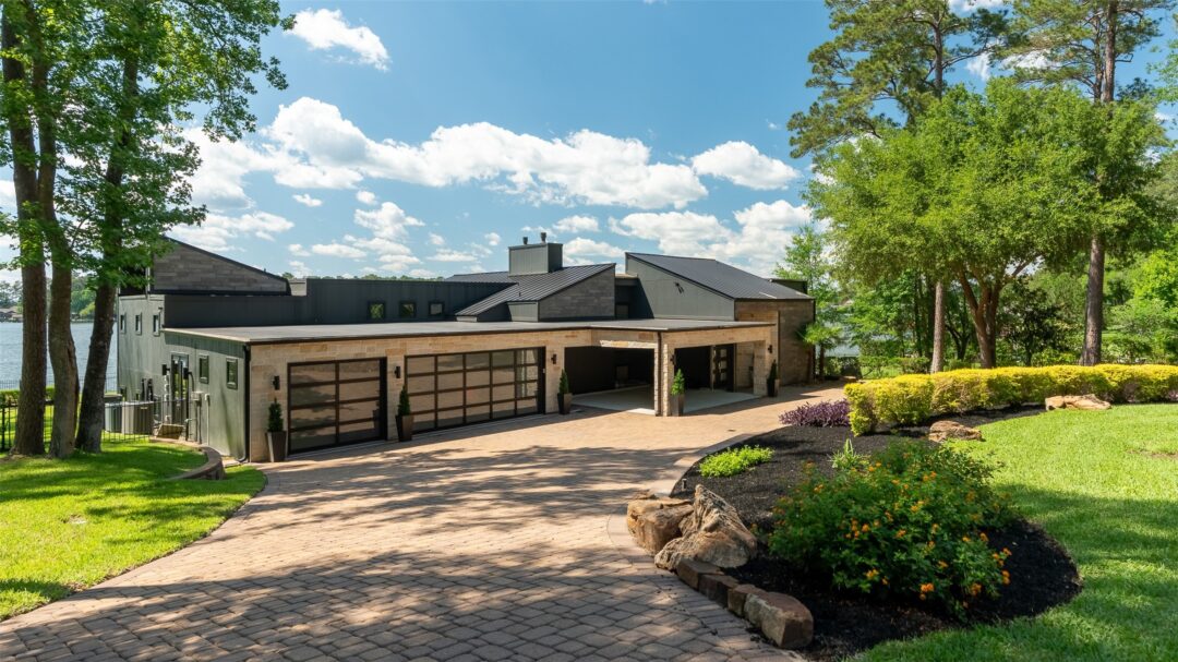 Modern Contemporary Home with Dark Metal Roof and Large Glass Garage Doors Set Along a Driveway with Landscaped Front Yard and Lake in the Background