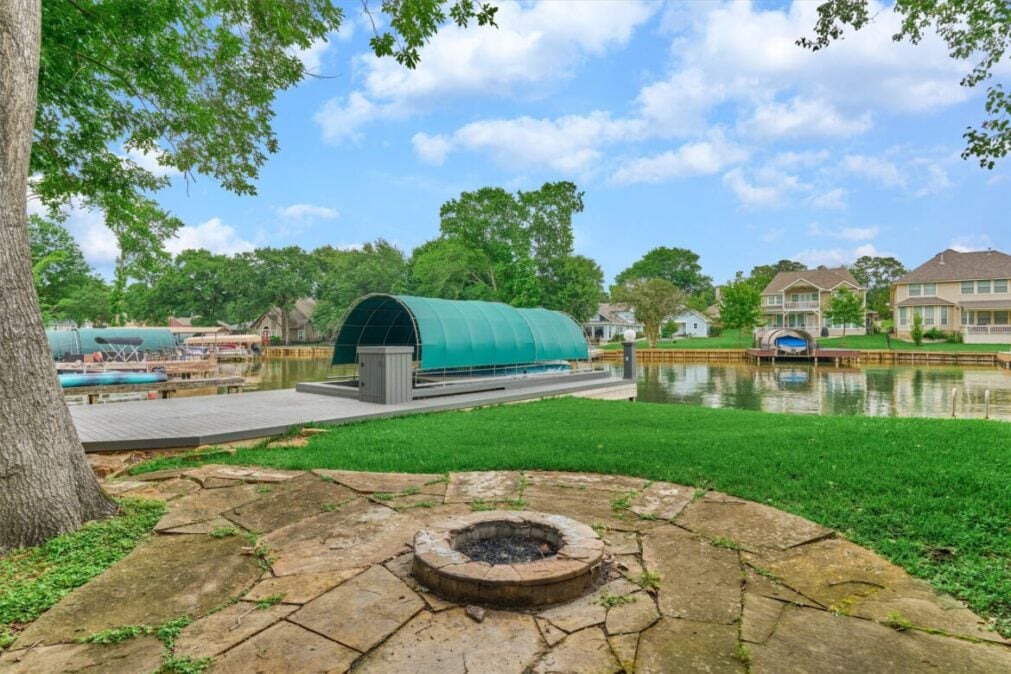 Lakeside Backyard with a Circular Stone Fire Pit in the Foreground a Deck and Teal Boat Shelter by Calm Water