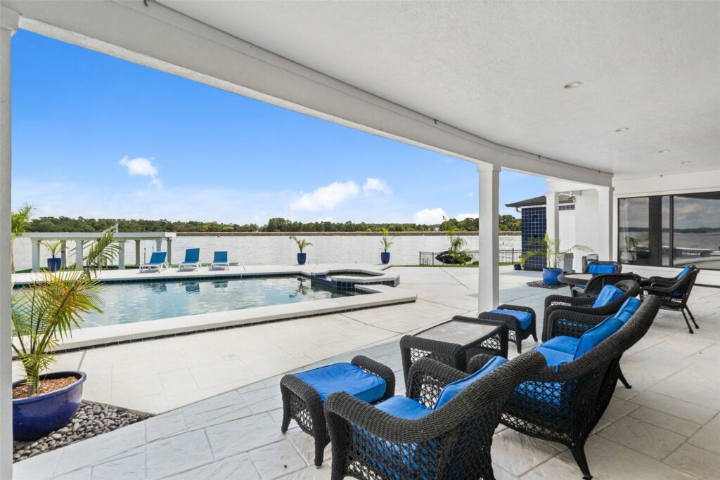 | the mckellar group Covered Patio with Wicker Seating and Bright Blue Cushions Overlooking a Rectangular Pool and a Calm Waterfront Lagoon