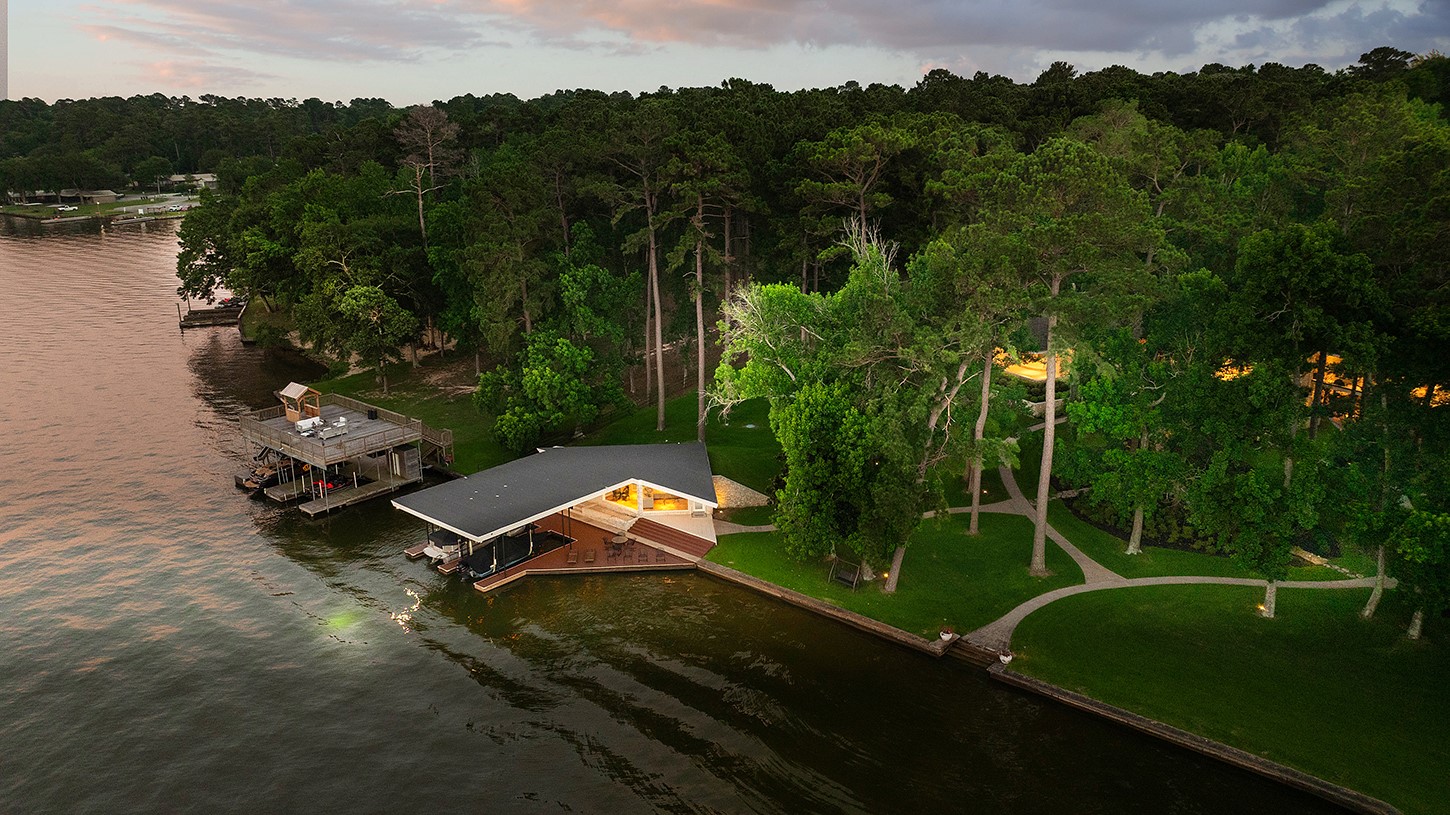 a Lakeside House with a Wraparound Deck by a Calm Shoreline Surrounded by Tall Trees at Dusk