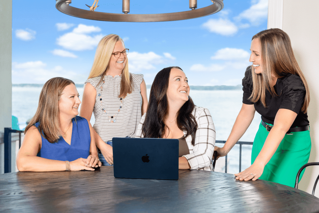 | the mckellar group Four Women Around a Wooden Table Outdoors Smiling at a Dark Blue Apple Laptop with the Sea and Blue Sky Behind Them