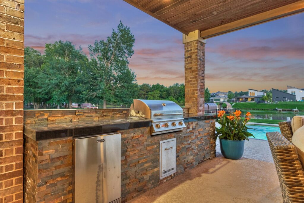 | the mckellar group Outdoor Kitchen with a Built in Stainless Grill and Stone Counter Under a Wooden Roof Facing a Pool at Sunset