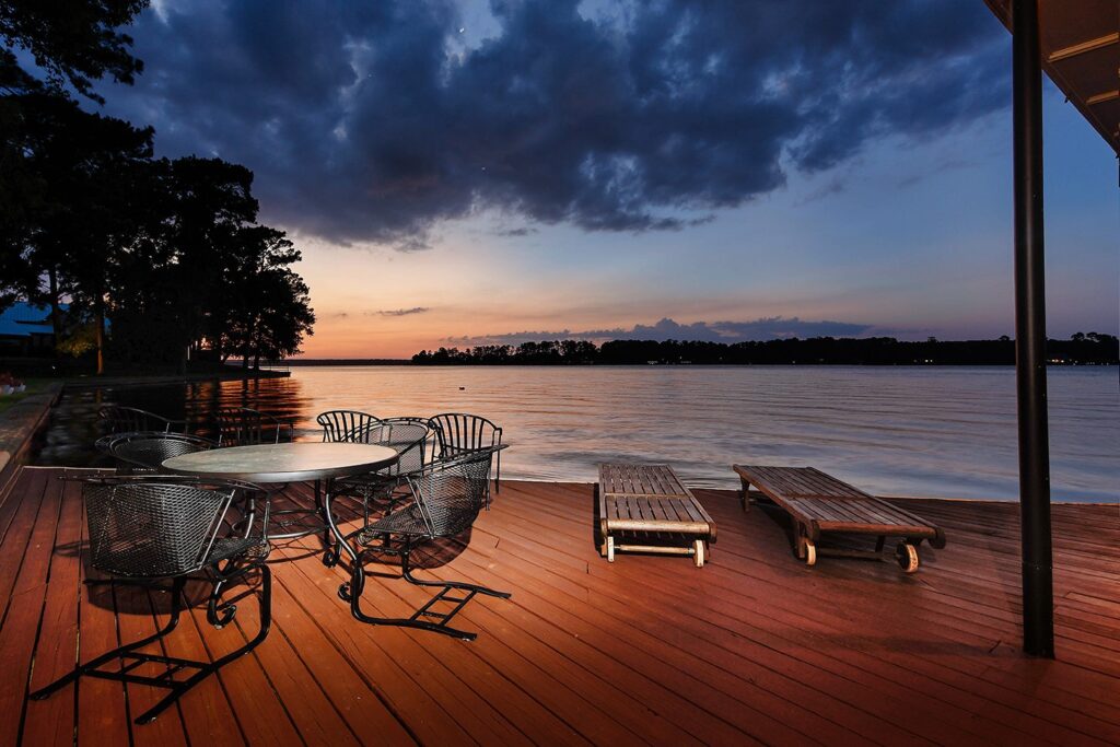 | the mckellar group Wooden Lakeside Deck at Sunset with a Round Metal Table and Iron Chairs and Two Lounge Chairs Overlooking Calm Water