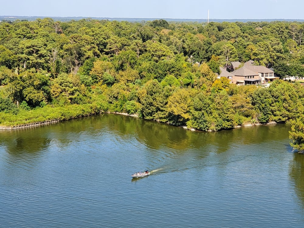 | the mckellar group Aerial View of a Wooded Shoreline with Calm Water and a Small Motorboat Traveling Across the Center Houses Peek Through Trees on the Right